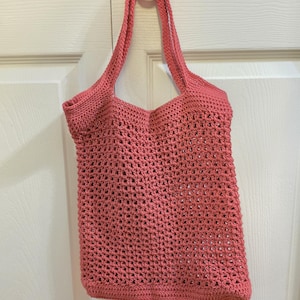 Può includere: Una borsa tote all'uncinetto color corallo caratterizzata da un motivo a rete traforata su tutto il corpo e due tracolle integrate. La borsa è realizzata con una consistenza del filato uniforme e mantiene una forma rettangolare con una base solida.