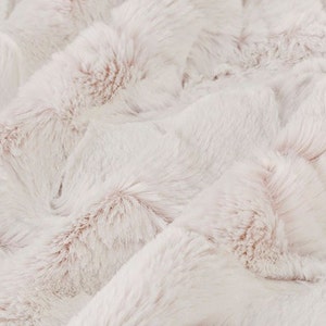 Puede incluir: Tela de piel sintética suave y esponjosa de color blanco con un sutil tono rosado. La piel está dispuesta en un patrón ondulado, creando una textura lujosa y acogedora.