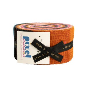 Pode incluir: Um rolo de tiras de tecido em tons de laranja, castanho e rosa. O tecido é preso com uma fita preta e uma etiqueta branca com texto. O rolo é rotulado como "Pixel" e "Ruby's Star Society".