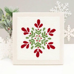 Peut inclure: Une toile carrée blanc cassé présente un flocon de neige brodé. Le motif du flocon de neige est composé de détails rouges et verts, avec de petits points rouges au centre. L'œuvre est encadrée d'une bordure neutre, évoquant un thème hivernal festif.