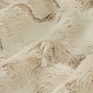 May include: Close-up of a soft, plush beige faux fur fabric. The fabric has a subtle wave pattern and a luxurious feel.
