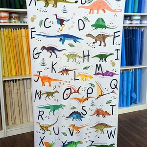 May include: A white fabric with a colorful alphabet printed on it. Each letter is paired with a dinosaur illustration. The dinosaurs are in various colors, including green, orange, purple, and blue. The fabric has a white background with black outlines around the letters and dinosaurs.