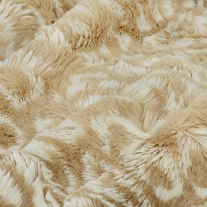 May include: A close-up of a soft, plush fabric with a light brown and white color scheme. The fabric has a fuzzy texture and a subtle pattern.