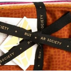 May include: A set of fabric squares in various shades of orange, pink, and red, tied together with a black ribbon. The ribbon has the text "Rue Star Society" printed on it.