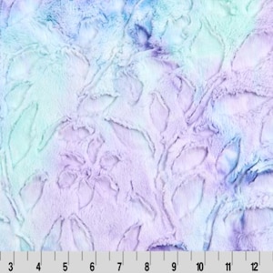 May include: A plush fabric with a floral design in shades of lavender, blue, and mint green. The fabric has a soft, textured appearance, with a ruler at the bottom showing measurements in inches.