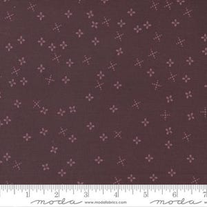 May include: A dark burgundy fabric with a repeating pattern of small, light pink floral and cross-shaped designs. The fabric has a textured appearance and is suitable for quilting or other craft projects. A ruler is at the bottom.