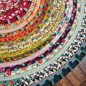 May include: A colorful round rug made from strips of fabric sewn together in a circular pattern. The rug has a variety of floral and geometric patterns in shades of red, orange, yellow, green, blue, and purple.