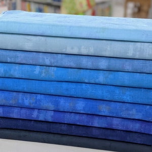 May include: A stack of ten fabric squares in shades of blue, ranging from light blue to dark blue. The fabric has a subtle texture and a slightly faded appearance.