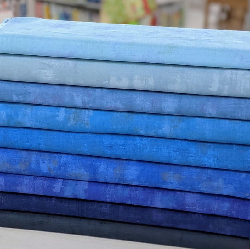 May include: A stack of ten fabric squares in shades of blue, ranging from light blue to dark blue. The fabric has a subtle texture and a slightly faded appearance.