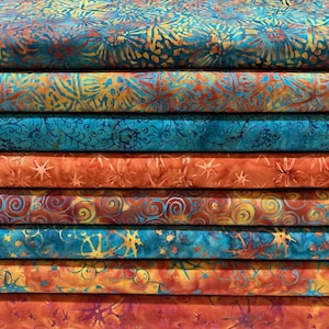 May include: A stack of eight folded fabric bolts featuring batik patterns in vibrant shades of turquoise, teal, orange, and yellow. The textiles display various motifs including starbursts, swirling lines, and organic shapes across the layered surfaces.