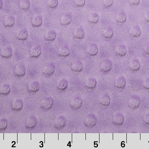 Puede incluir: Primer plano de una tela minky lavanda con un patrón de puntos en relieve. El material suave y afelpado tiene una superficie texturizada con protuberancias redondeadas y espaciadas uniformemente. La tela se mide con una regla en centímetros.