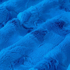 May include: Close-up of a soft, blue faux fur fabric with a wavy texture. The fabric is plush and has a luxurious feel.