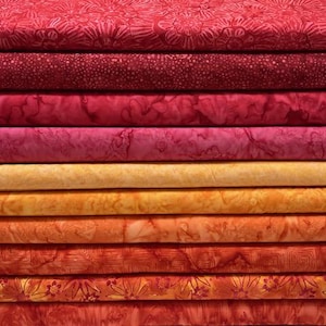 Puede incluir: Una pila de diez rollos de tela doblados dispuestos en un degradado de rojo intenso a naranja brillante. Cada pieza de tela presenta un patrón único estilo batik, incluyendo motivos florales, líneas geométricas y texturas moteadas.