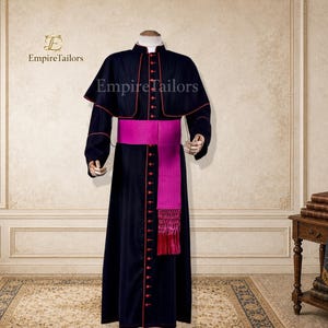 May include: A black cassock with a short cape, featuring red piping along the edges and a vertical row of red buttons. A wide, bright magenta sash with a textured finish and fringed ends is worn around the waist. The text EmpireTailors is visible.
