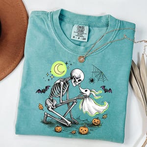 May include: A teal short-sleeved t-shirt with a graphic print of a skeleton kneeling before a ghost dog. The design includes a yellow crescent moon, stars, bats, a spider web, and three small pumpkins. The shirt features a Comfort Colors brand label.