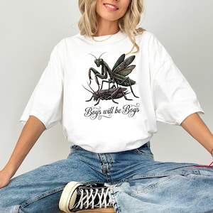 May include: White t-shirt featuring a detailed illustration of a praying mantis and a cricket, with the text "Boys will be Boys" in a decorative font. The graphic is in shades of green and black.