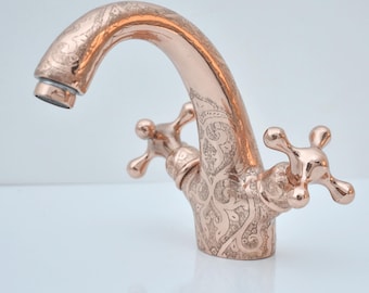 May include: A copper-colored faucet with an ornate, engraved floral pattern. The faucet features a curved spout and two cross-shaped handles. The design is vintage and elegant, suitable for a bathroom or kitchen.