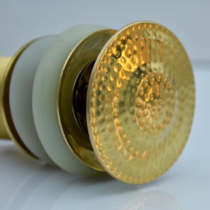May include: Close-up of a decorative bottle stopper. The stopper features a gold-colored, hammered metal disc, a light green ring, and a gold-colored base. The design is elegant and detailed.