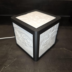 May include: A white cube-shaped lamp with a black frame. The sides of the lamp feature a detailed, textured design. The lamp is illuminated, and a white cord extends from the base.