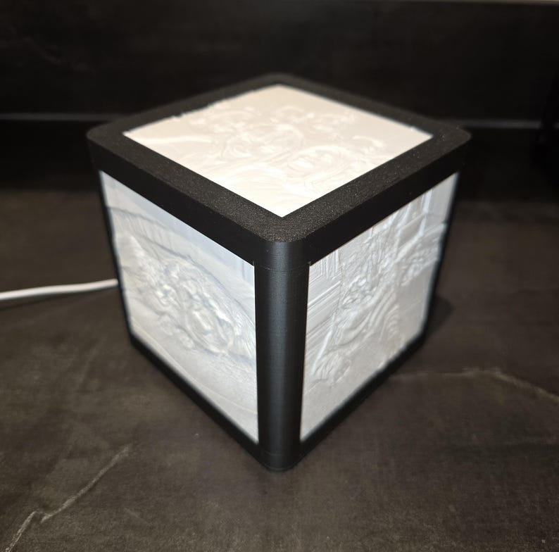 May include: A white cube-shaped lamp with a black frame. The sides of the lamp feature a detailed, textured design. The lamp is illuminated, and a white cord extends from the base.