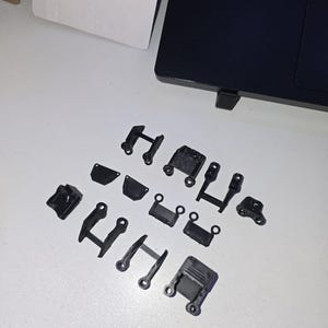 May include: A collection of small, black, 3D-printed plastic parts, likely for a mechanical or electronic device, arranged on a white surface. The parts vary in shape, including brackets, hinges, and connectors, with a matte finish.