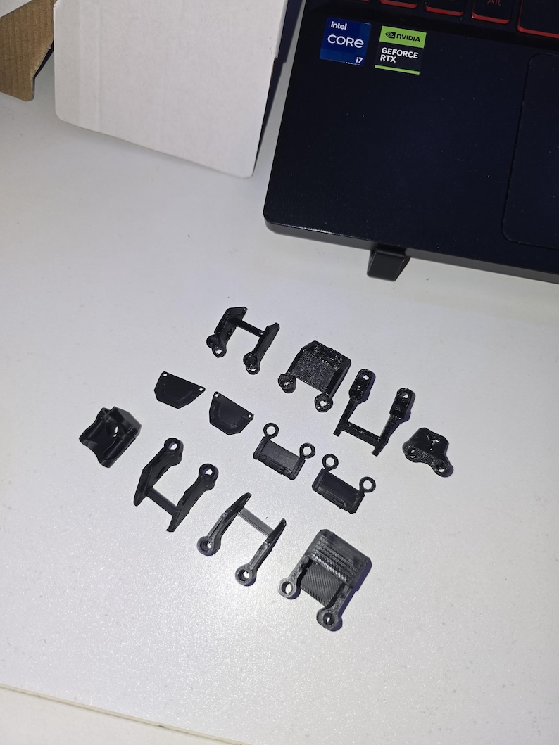 May include: A collection of small, black, 3D-printed plastic parts, likely for a mechanical or electronic device, arranged on a white surface. The parts vary in shape, including brackets, hinges, and connectors, with a matte finish.