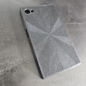 May include: A black phone case with a textured, geometric design. The case has a camera hole and a small opening on the side. The case is rectangular and appears to be made of a rigid material.