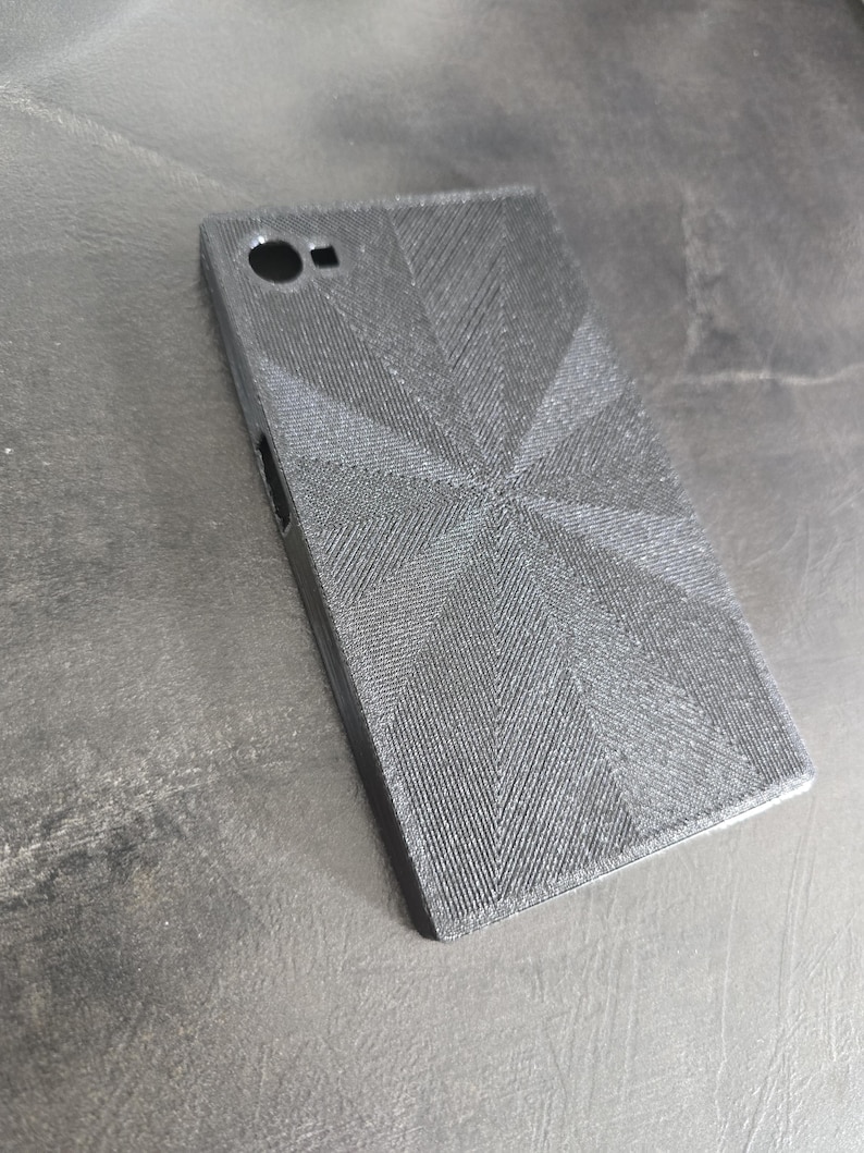 May include: A black phone case with a textured, geometric design. The case has a camera hole and a small opening on the side. The case is rectangular and appears to be made of a rigid material.