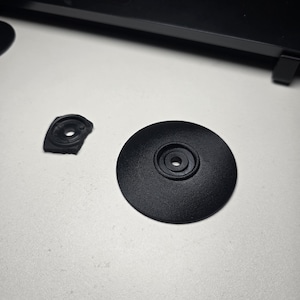 May include: Two black plastic components, one circular and one irregularly shaped, are displayed on a white surface. The circular piece has a central hole and a textured surface. The other piece is broken.