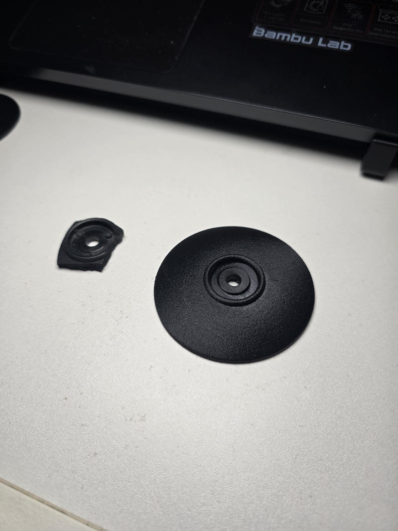 May include: Two black plastic components, one circular and one irregularly shaped, are displayed on a white surface. The circular piece has a central hole and a textured surface. The other piece is broken.