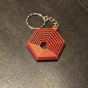 May include: Red geometric keychain with a silver chain and keyring. The keychain has a layered hexagonal design, creating a three-dimensional effect. The keychain is made of a solid material and is set against a dark background.