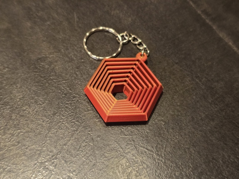 May include: Red geometric keychain with a silver chain and keyring. The keychain has a layered hexagonal design, creating a three-dimensional effect. The keychain is made of a solid material and is set against a dark background.