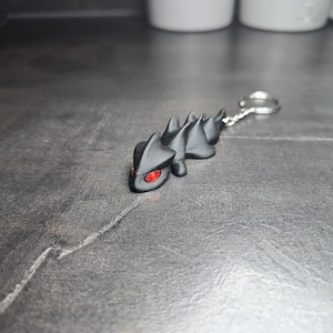 May include: A black keychain featuring a stylized lizard with red eyes. The lizard has a spiky back and is attached to a silver key ring. The keychain is made of a dark material and rests on a textured gray surface.