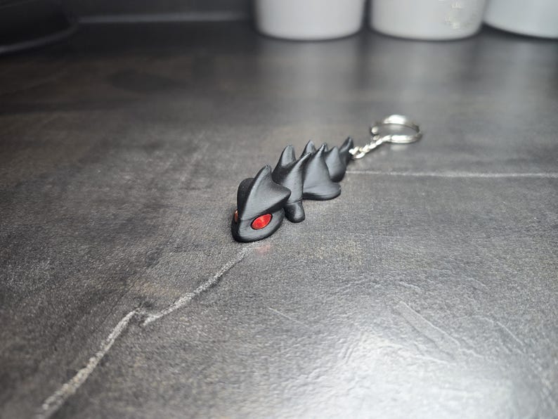 May include: A black keychain featuring a stylized lizard with red eyes. The lizard has a spiky back and is attached to a silver key ring. The keychain is made of a dark material and rests on a textured gray surface.