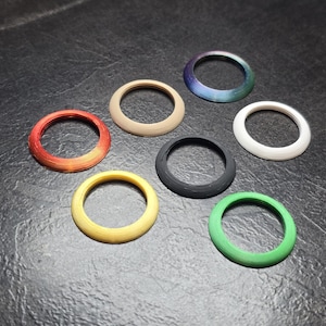 May include: A collection of seven circular rings in various colors including orange, beige, iridescent blue-purple, white, black, yellow, and green. Each ring features a beveled edge and a smooth, uniform finish, arranged in a scattered pattern.