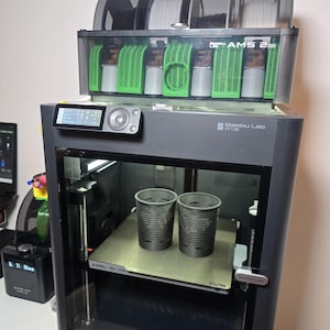 以下が含まれることがあります： ダークグレーのBambu Lab P1S 3Dプリンター。上部には4つのフィラメントスプールを収納した透明なBambu Lab AMSユニットが設置されている。プリンター内部のプリントベッド上には、格子模様のダークグレーの円筒形容器が2つ置かれている。