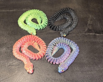 May include: Four articulated snake figurines in green, black, orange, and a blue-purple gradient. Each snake features a textured, scale-like surface, a coiled body, and small, distinct eyes. The figures are arranged in a two-by-two grid formation.