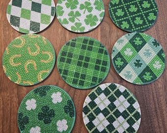 May include: A collection of eight round coasters with various St. Patrick's Day-themed designs. The coasters feature patterns like shamrocks, plaid, and horseshoes in shades of green and white. The coasters are arranged on a wooden surface.