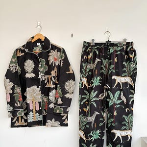 Puede incluir: Conjunto de pijama negro con estampado de jungla con cebras, leopardos y plantas tropicales. La parte superior de manga larga tiene ribetes y botones blancos. Los pantalones tienen cintura con cordón y ribete a juego.