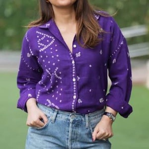 Puede incluir: Una camisa morada de manga larga con un estampado de puntos blancos, combinada con vaqueros azul claro. La camisa tiene cuello y puños abotonados. La persona lleva un reloj. La camisa es de tejido suave.