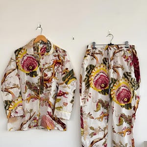 Könnte beinhalten: Ein zweiteiliger Schlafanzug mit einem Hemd mit Knöpfen und einer Hose. Das Set ist weiß mit einem Blumenmuster in Rosa, Gelb, Grün und Braun. Das Hemd hat lange Ärmel und gelbe Paspeln. Die Hose hat einen elastischen Bund.