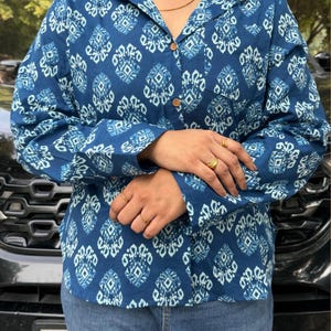 May include: A long-sleeved, button-down shirt in a deep blue hue, adorned with a repeating white geometric pattern. The shirt features a classic collar and a single chest pocket. The sleeves are cuffed, and the shirt is paired with blue jeans.