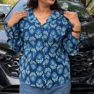 May include: A blue button-down shirt with a white geometric pattern. The shirt has long sleeves and wooden buttons. The shirt is paired with blue jeans. The shirt is a collared shirt.
