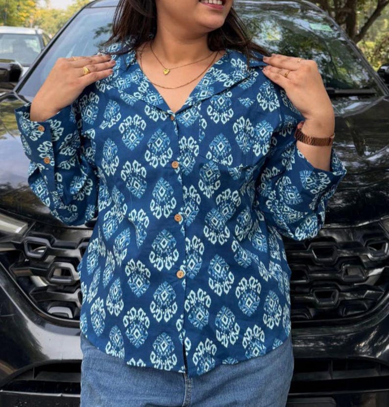 May include: A blue button-down shirt with a white geometric pattern. The shirt has long sleeves and wooden buttons. The shirt is paired with blue jeans. The shirt is a collared shirt.
