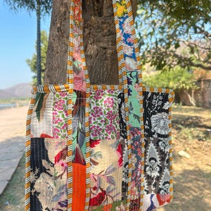 May include: A patchwork tote bag with a variety of floral patterns in shades of pink, red, blue, and black. The bag has a rectangular shape with a sturdy base and long straps. The straps and edges are trimmed with a yellow and orange striped fabric.