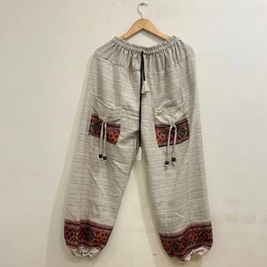 Könnte beinhalten: Hellgraue Hose mit zwei Vordertaschen und Zierleisten. Die Taschen und Manschetten weisen ein farbenfrohes geometrisches Muster in Rot, Braun und Creme auf. Die Hose hat einen Kordelzugbund und hängt an einem Holzbügel.