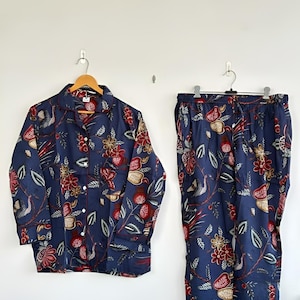 May include: Navy blue pajama set with a floral pattern in red, gold, and green. The long-sleeved top has a button-down front and a notched collar. The matching pants have a drawstring waist. The set is hanging on a white wall.
