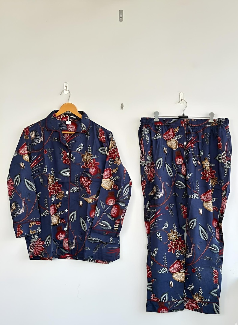 May include: Navy blue pajama set with a floral pattern in red, gold, and green. The long-sleeved top has a button-down front and a notched collar. The matching pants have a drawstring waist. The set is hanging on a white wall.