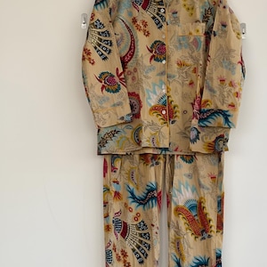 May include: A beige pajama set with a colorful floral and paisley pattern. The long-sleeved top has a button-down front and a collar. The pants are long and have a drawstring waist. The set is hanging on a wooden hanger.