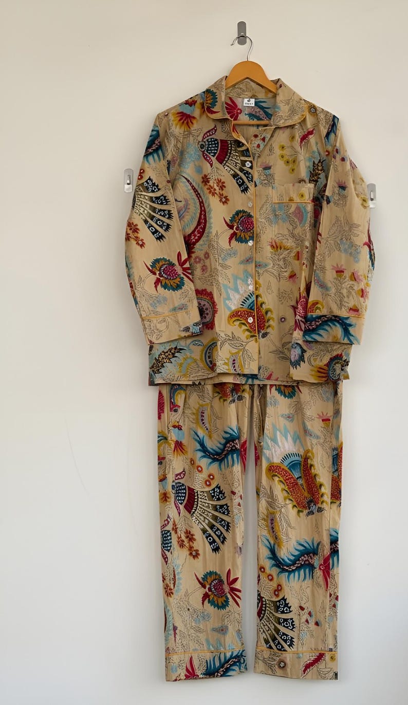 May include: A beige pajama set with a colorful floral and paisley pattern. The long-sleeved top has a button-down front and a collar. The pants are long and have a drawstring waist. The set is hanging on a wooden hanger.